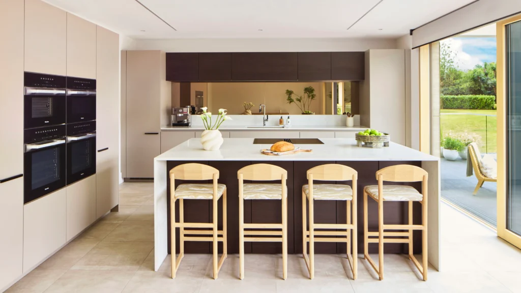 Modern open-plan kitchen with integrated Miele appliances, central island with blonde wood stools, and natural light flooding through large windows