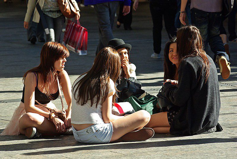 A group of four young women sitting on the ground in a public square engaged in conversation while pedestrians walk past in the background.
