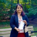 Rashmi Prakash, UBC biomedical engineering alumna, standing outdoors holding a compostable menstrual pad product while wearing a navy blazer over a floral dress.
