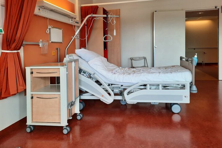 Empty hospital room with an adjustable medical bed, bedside cabinet, IV stand, and medical equipment against orange walls.