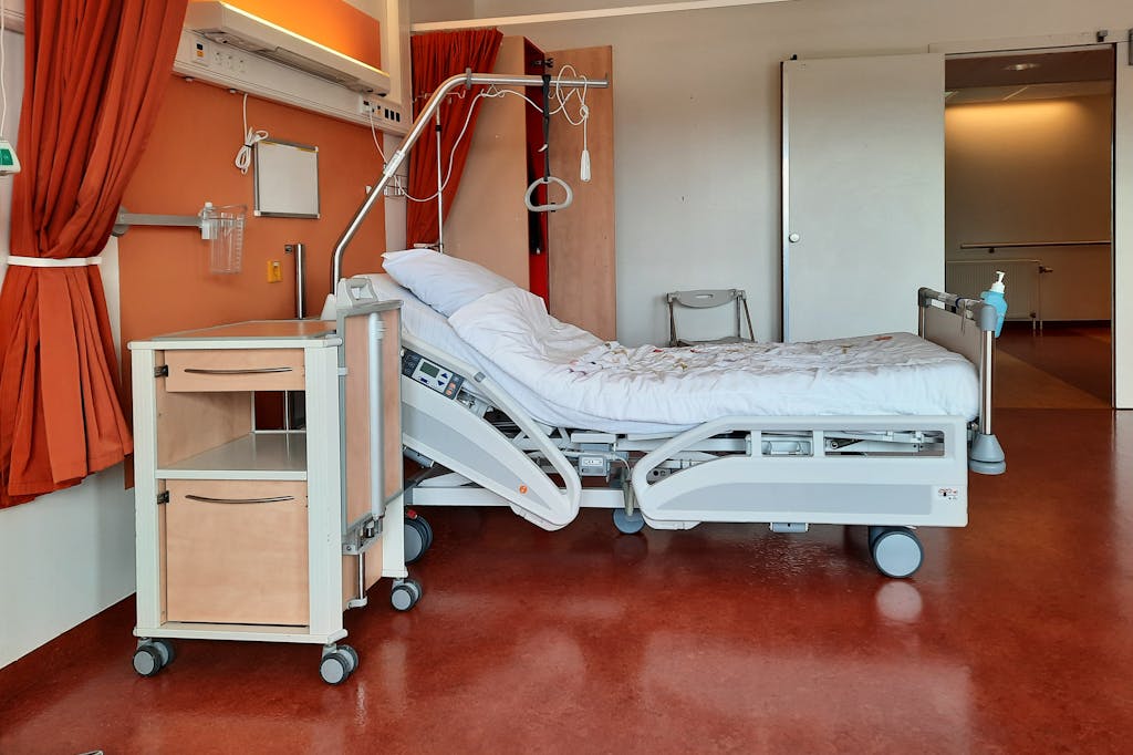 Empty hospital room with an adjustable medical bed, bedside cabinet, IV stand, and medical equipment against orange walls.