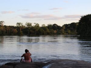 Indigenous person sitting on a rock overlooking a river in the Amazon rainforest at sunset, surrounded by dense forest vegetation.