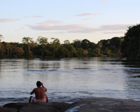 Indigenous person sitting on a rock overlooking a river in the Amazon rainforest at sunset, surrounded by dense forest vegetation.