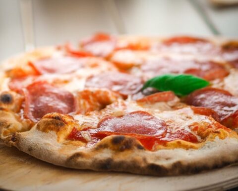 Close-up of a wood-fired pepperoni pizza with melted cheese, slices of pepperoni, and a single green basil leaf on a wooden surface.