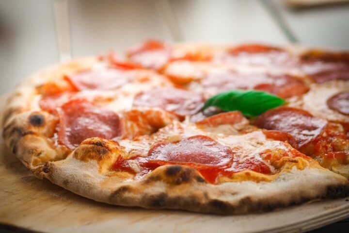 Close-up of a wood-fired pepperoni pizza with melted cheese, slices of pepperoni, and a single green basil leaf on a wooden surface.