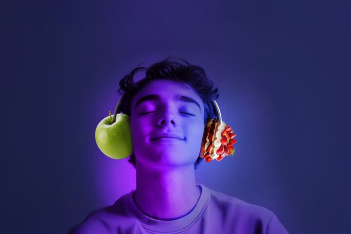 Person with closed eyes wearing headphones, with a green apple positioned at one ear and strawberries at the other ear, against a purple-blue background.