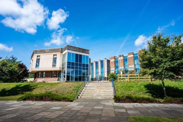 Modern university building at the University of Kent campus featuring glass facades and stone architecture with steps leading to the entrance, surrounded by green lawns and trees under a blue sky with white clouds.