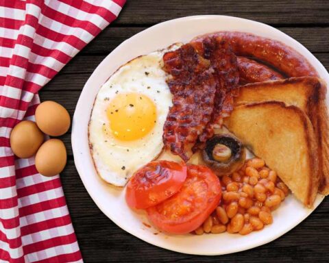 Traditional English breakfast with fried egg, bacon, beans, toast, and tomato served on a white plate next to brown eggs on a red and white striped cloth
