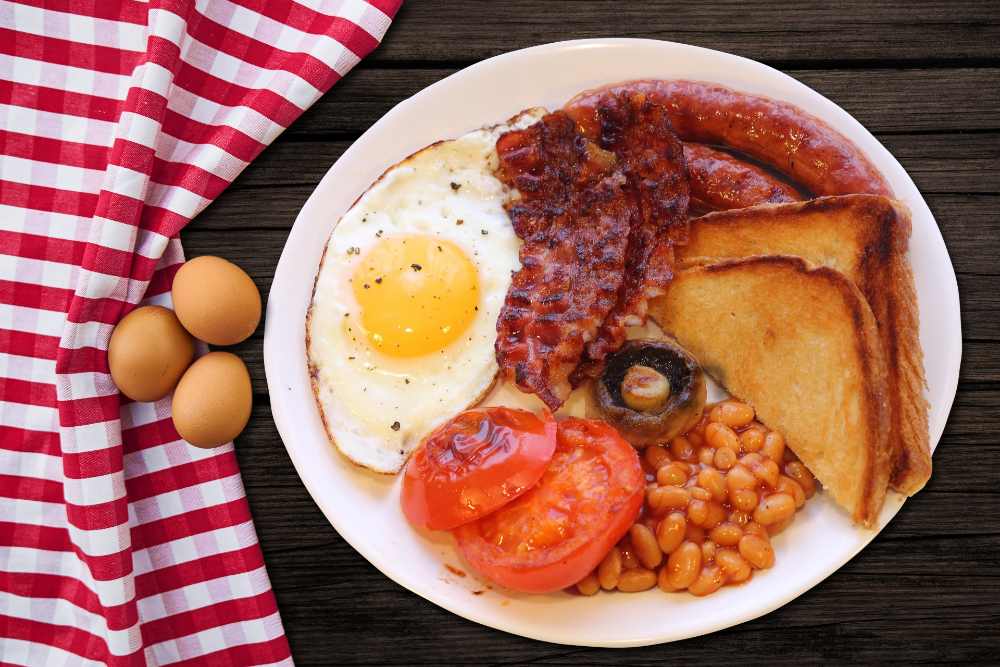 Traditional English breakfast with fried egg, bacon, beans, toast, and tomato served on a white plate next to brown eggs on a red and white striped cloth