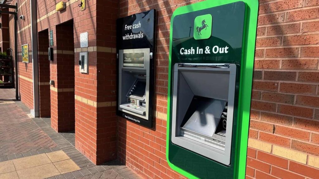 A Lloyds Bank ATM with green trim showing "Cash In & Out" signage mounted on a brick wall exterior of a bank branch.