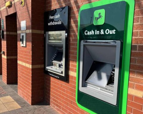 A Lloyds Bank ATM with green trim showing "Cash In & Out" signage mounted on a brick wall exterior of a bank branch.