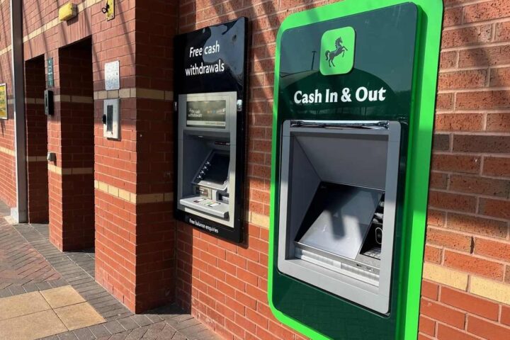 A Lloyds Bank ATM with green trim showing "Cash In & Out" signage mounted on a brick wall exterior of a bank branch.