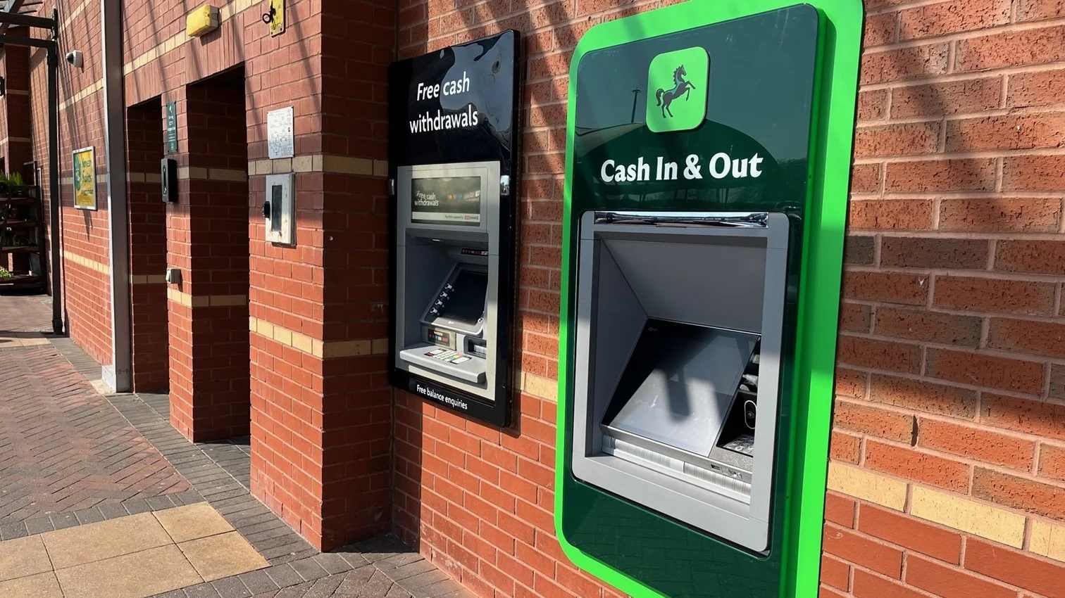 A Lloyds Bank ATM with green trim showing "Cash In & Out" signage mounted on a brick wall exterior of a bank branch.