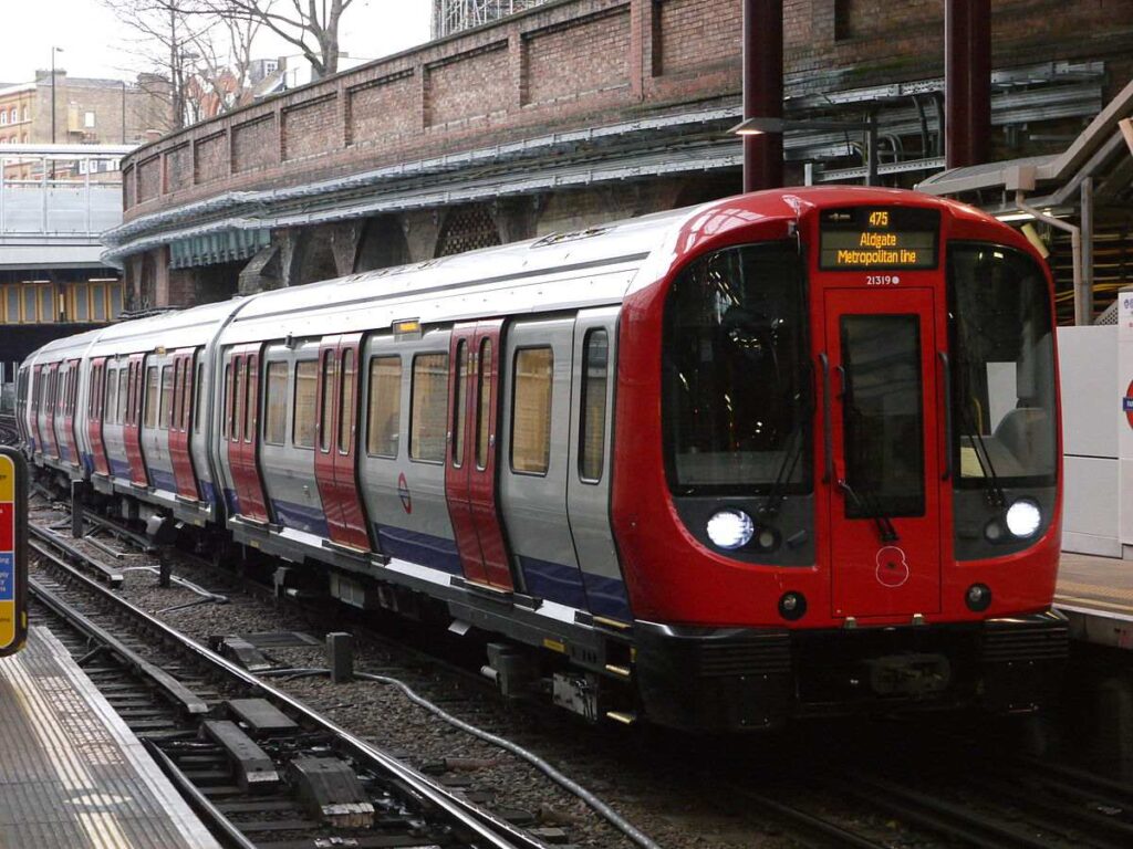 A London Underground train with distinctive red livery stopped at a platform in a brick-walled station with metal support columns.