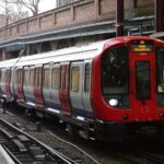 A London Underground train with distinctive red livery stopped at a platform in a brick-walled station with metal support columns.