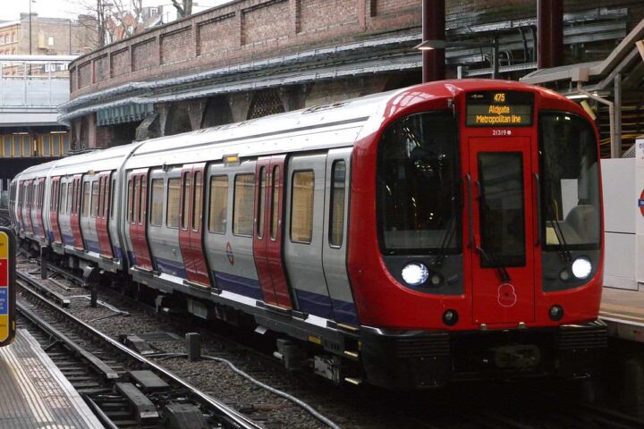 A London Underground train with distinctive red livery stopped at a platform in a brick-walled station with metal support columns.