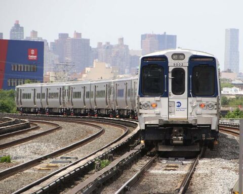 A Long Island Rail Road M9 train bound for Penn Station traveling on curved tracks as it leaves Woodside, New York, with city buildings in the background.