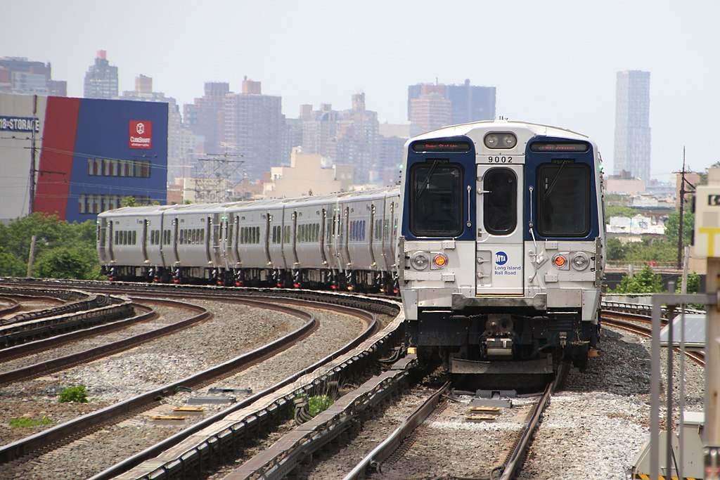 A Long Island Rail Road M9 train bound for Penn Station traveling on curved tracks as it leaves Woodside, New York, with city buildings in the background.