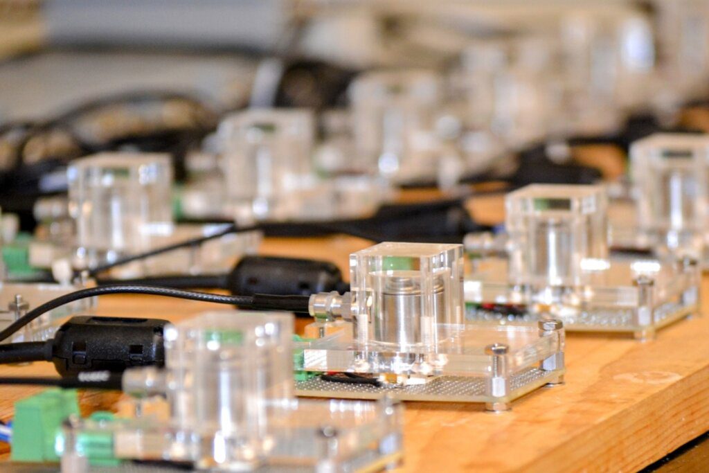 Laboratory setup showing transparent battery test cells arranged on a wooden table with connected sensors and monitoring equipment for acoustic emissions research.