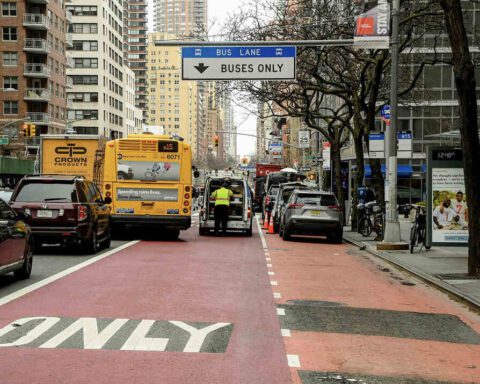 A New York City bus traveling in a red-painted bus lane with "BUSES ONLY" marked on the pavement and a blue overhead sign indicating "BUS LANE" while other vehicles are visible in adjacent lanes.
