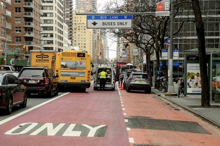 A New York City bus traveling in a red-painted bus lane with "BUSES ONLY" marked on the pavement and a blue overhead sign indicating "BUS LANE" while other vehicles are visible in adjacent lanes.