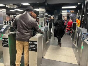 Passengers using MTA turnstiles at a subway station, with one person tapping their MetroCard to enter while others walk through the fare gates. A person in a wheelchair is visible being assisted by MTA staff near an elevator sign.
