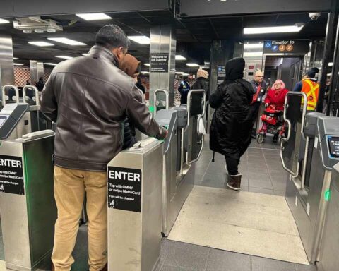 Passengers using MTA turnstiles at a subway station, with one person tapping their MetroCard to enter while others walk through the fare gates. A person in a wheelchair is visible being assisted by MTA staff near an elevator sign.