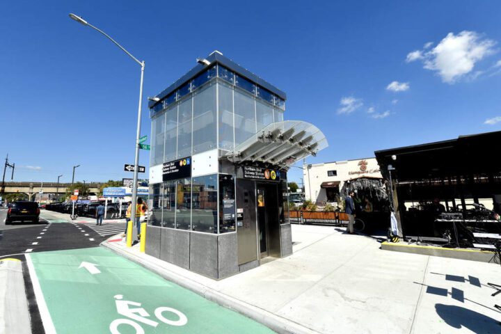 Glass-enclosed elevator structure at Northern Boulevard subway station with bike lane visible in foreground and station entrance nearby