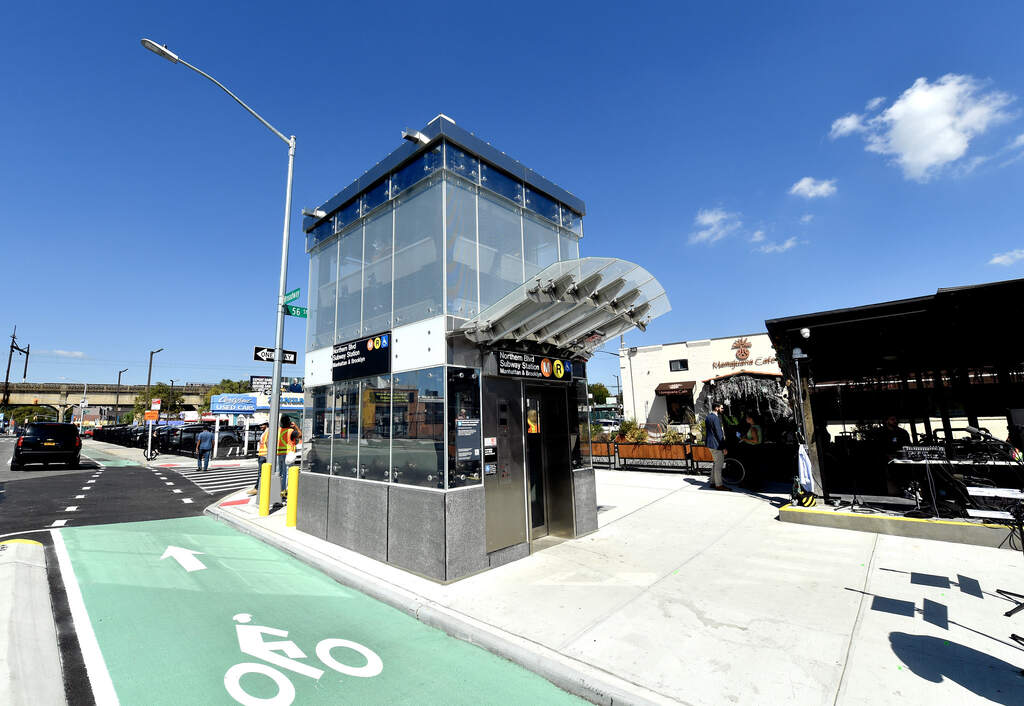 Glass-enclosed elevator structure at Northern Boulevard subway station with bike lane visible in foreground and station entrance nearby