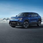 Blue Maruti e VITARA electric SUV positioned on dark ground with snowy mountains in the background, showcasing its modern crossover design with distinctive front lighting.
