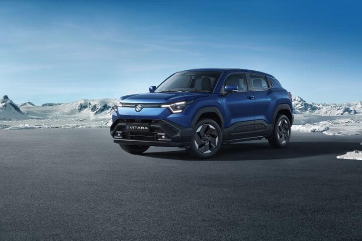 Blue Maruti e VITARA electric SUV positioned on dark ground with snowy mountains in the background, showcasing its modern crossover design with distinctive front lighting.