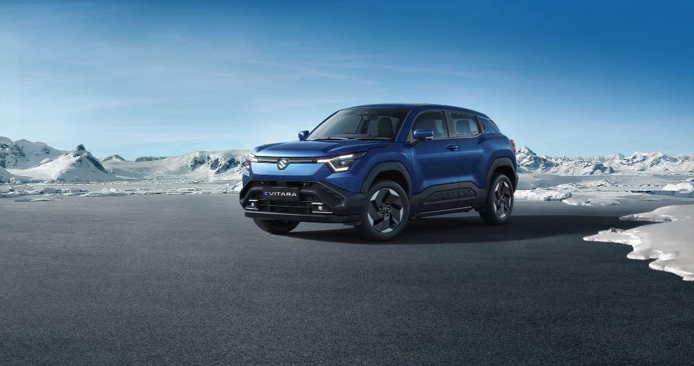 Blue Maruti e VITARA electric SUV positioned on dark ground with snowy mountains in the background, showcasing its modern crossover design with distinctive front lighting.