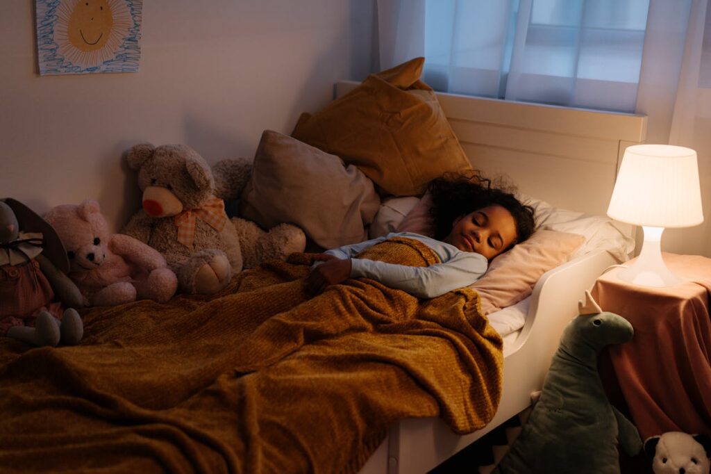 A young child with curly hair sleeping peacefully in bed with stuffed animals, bedside lamp on, covered with a mustard yellow blanket.