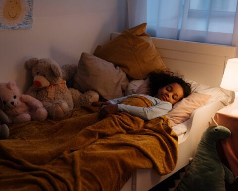 A young child with curly hair sleeping peacefully in bed with stuffed animals, bedside lamp on, covered with a mustard yellow blanket.