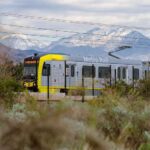 Metro Rail light rail train with yellow front section traveling through brushy terrain with snow-covered San Gabriel Mountains rising dramatically in the background.