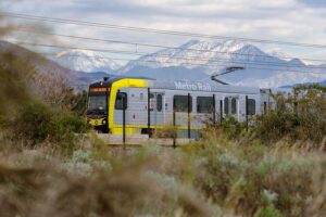 Metro Rail light rail train with yellow front section traveling through brushy terrain with snow-covered San Gabriel Mountains rising dramatically in the background.
