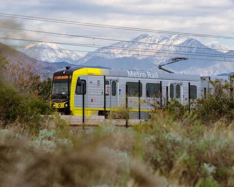 Metro Rail light rail train with yellow front section traveling through brushy terrain with snow-covered San Gabriel Mountains rising dramatically in the background.