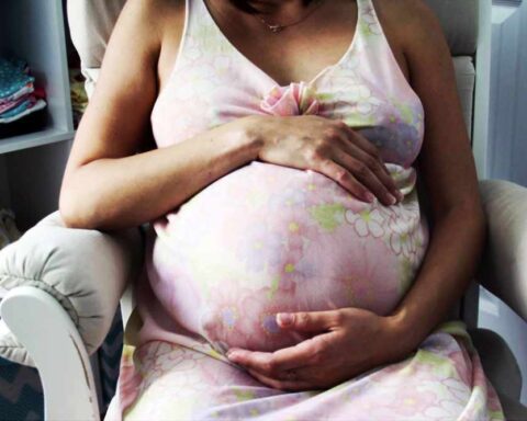 Pregnant woman in pink dress sitting in nursery chair holding her belly with both hands
