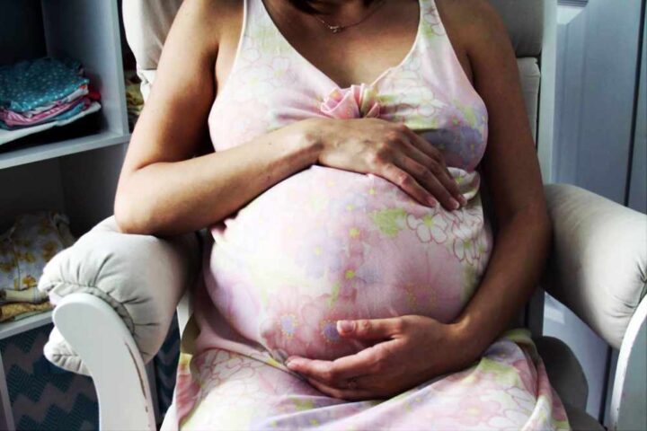 Pregnant woman in pink dress sitting in nursery chair holding her belly with both hands