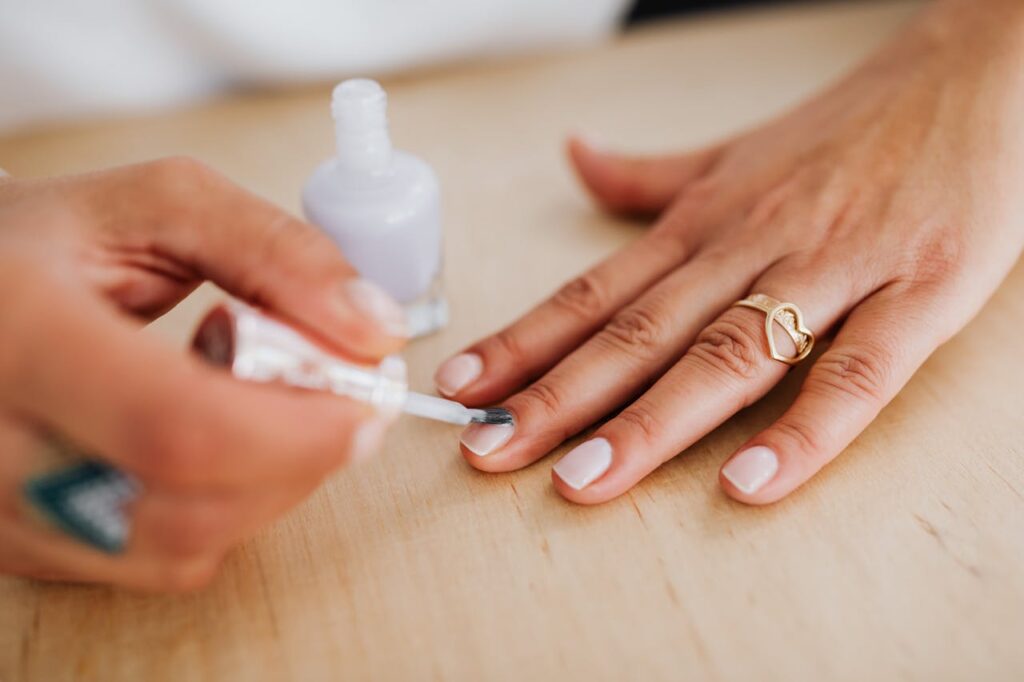 Close-up of a manicurist applying white nail polish to a woman's hand wearing a gold heart-shaped ring on a wooden table surface.