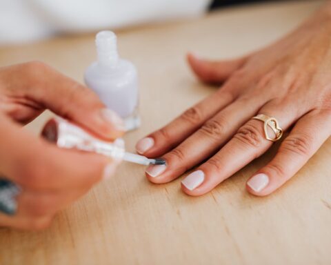 Close-up of a manicurist applying white nail polish to a woman's hand wearing a gold heart-shaped ring on a wooden table surface.