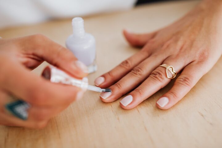 Close-up of a manicurist applying white nail polish to a woman's hand wearing a gold heart-shaped ring on a wooden table surface.