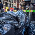 Black garbage bags piled beside a metal trash can on a New York City street in the Theatre District, with pedestrians and shops in the background.