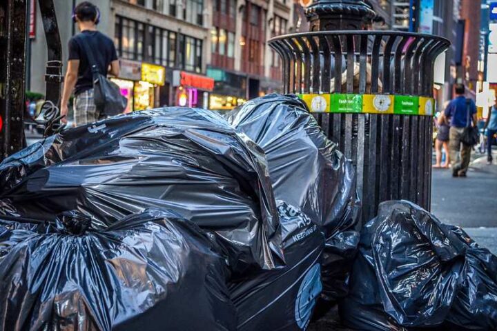 Black garbage bags piled beside a metal trash can on a New York City street in the Theatre District, with pedestrians and shops in the background.