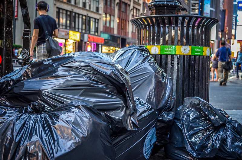 Black garbage bags piled beside a metal trash can on a New York City street in the Theatre District, with pedestrians and shops in the background.