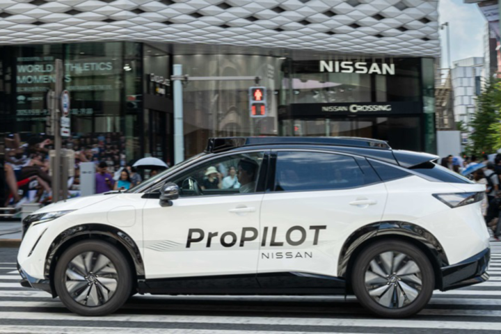A white Nissan Ariya electric vehicle with "ProPILOT" written on its side drives through a busy Tokyo intersection with pedestrians and buildings in the background.