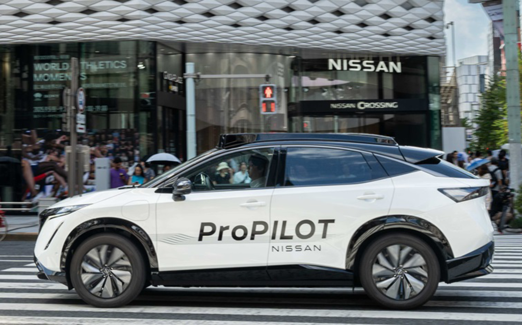 A white Nissan Ariya electric vehicle with "ProPILOT" written on its side drives through a busy Tokyo intersection with pedestrians and buildings in the background.