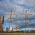Overhead power lines and transmission towers stand against a cloudy sky, with residential buildings and industrial structures in the background.