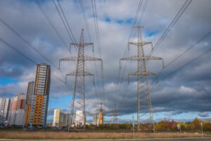 Overhead power lines and transmission towers stand against a cloudy sky, with residential buildings and industrial structures in the background.
