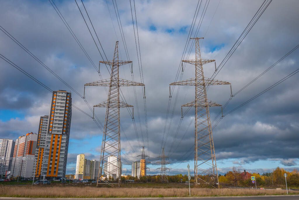 Overhead power lines and transmission towers stand against a cloudy sky, with residential buildings and industrial structures in the background.
